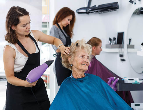 Professional female stylist making hairdo for senior woman in salon, using hair dryer 